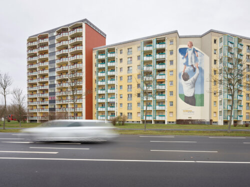 Toni Züchner, Neulandgewinner Runde 6. Hoyerswerda, von seinen Bewohner*innen auch liebevoll Hoywoj genannt, hat eine Alt- und eine Neustadt, zwischen denen die Schwarze Elster fließt. Was schon immer durch den Fluss getrennt war, klafft allerdings – durch den Abriss vieler Plattenbauten – immer stärker auseinander. Was die Stadt also braucht, ist: Verbindung.

Toni Züchner will sie schaffen. Der Plan, den der Projektleiter der Kulturfabrik e.V. gemeinsam mit einem breiten Bündnis anderer Akteur*innen vor Ort schmiedet, nennt sich „Der Grüne Saum“ – und ist ein 20 Kilometer langer Rundweg um Hoyerswerda, der Alt- und Neustadt umkreisen und wieder zu einem großen Ganzen machen soll. Züchner, der aus Hoyerswerda stammt und nach einem Kulturmanagement-Studium auch dorthin zurückgekehrt ist, will etwas dafür tun, dass man in seiner Heimatstadt „gut leben kann“, dass die Stimmung in der Bürgerschaft wieder optimistischer wird.

Den „Grünen Saum“ stellen sich Züchner und seine Projektpartner*innen als Kulturwanderweg und Naherholungspfad vor – mit einer attraktiv gestalteten grünen Mitte zwischen Alt- und Neustadt. Am Wegesrand sollen Wandernde, Spaziergänger*innen und Fahrradfahrer*innen Gastronomie und Spielplätze besuchen können. Den Pfad werden Grillplätze und eine Freilichtbühne säumen, Streuobstwiesen und temporäre Außen-Galerien, ein Naturlehrpfad sowie ein grünes Klassenzimmer.

Toni Züchner will das Projekt zunächst mit der Bevölkerung diskutieren und dann in enger Zusammenarbeit mit der Stadtverwaltung umsetzen. „Der kommunale Entwicklungsbeirat ist bereits gegründet, alles steht in den Startlöchern“, sagt er. „Ich bin sicher, dass das Projekt uns dabei helfen wird, den Strukturwandel zu bewältigen.“